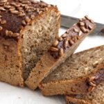 Square sunflower seed loaf sliced on rustic white surface.